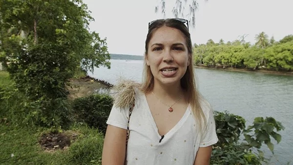 Woman talking to camera by river, lush greenery background
