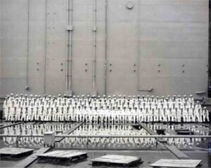 Sailors in uniform stand in formation before a large ship hull, possibly an ABSD.