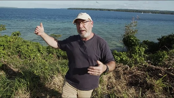 Man in a cap gestures towards a blue lake, discussing videos.