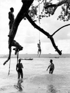 Aore: Boys on a rope swing over water, historical scene.