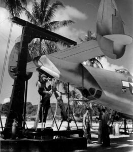 WWII aircraft maintenance: Mechanics service a bomber with a hoist in a tropical setting.