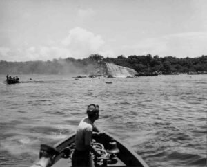 USS Coolidge wreck, listing heavily in shallow water with onlookers in boats.