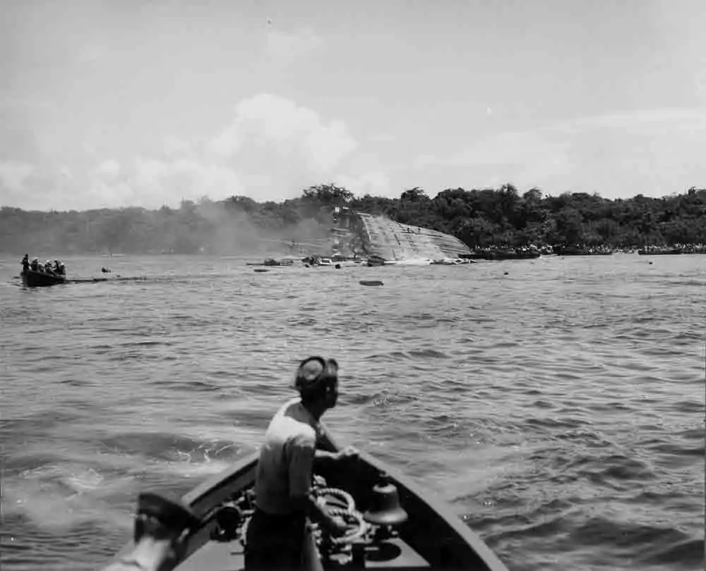 USS Coolidge wreck, listing heavily in shallow water with onlookers in boats.