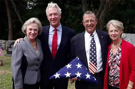 Four people in suits and dresses hold a folded American flag at a gravesite.