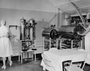 Nurses in a historical hospital room with autoclaves and a covered table.