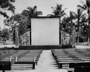 Outdoor cinema screen with rows of empty benches, palm trees in background