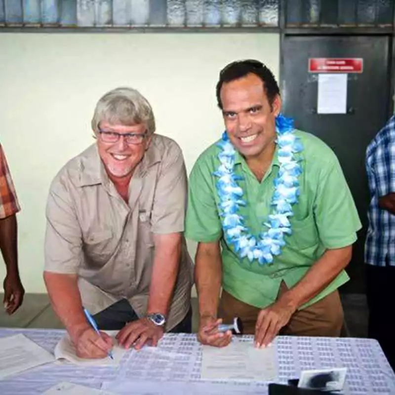 Two men signing documents, one wearing a lei, in a corporate setting.