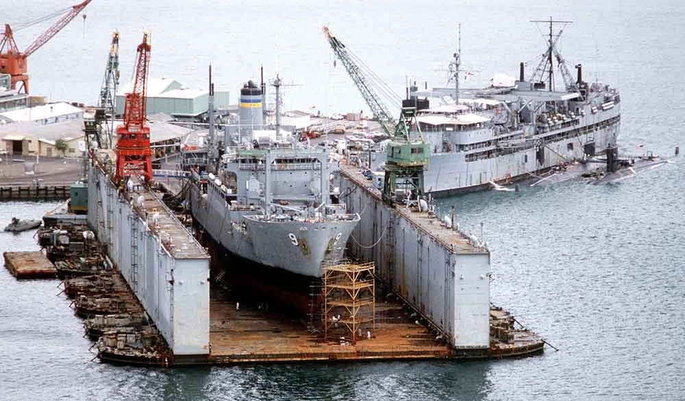 Naval ship in a floating dry dock, with cranes and other vessels nearby.