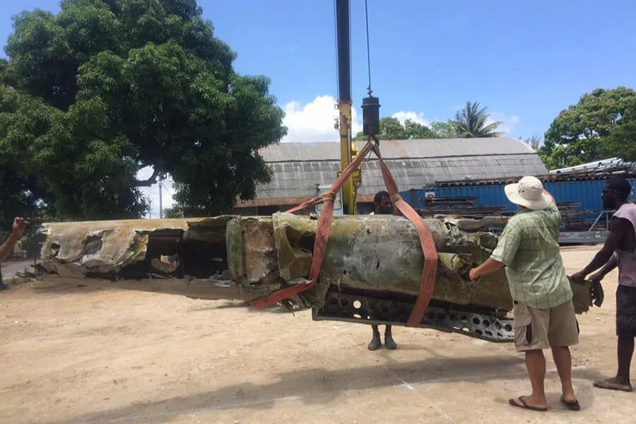Crane lifts salvaged aircraft wreckage in a dusty outdoor area.