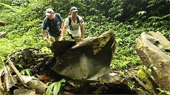 Two explorers examine wreckage in a dense jungle, possibly a plane crash site.