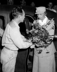 Nurse presents flowers to a man next to a car, historical Roosevelt context