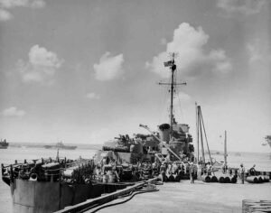 US Navy ship undergoing replenishment at sea, with sailors and supplies visible.