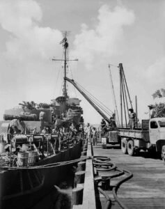 Naval replenishment: Sailors load supplies onto a warship from a dockside truck with a crane.