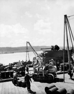 WWII replenishment: Crane lifts barrels onto a ship deck with soldiers.