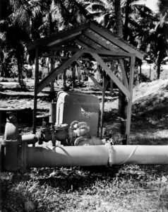 Vintage fuel pump and pipes under a wooden shelter, surrounded by palm trees.