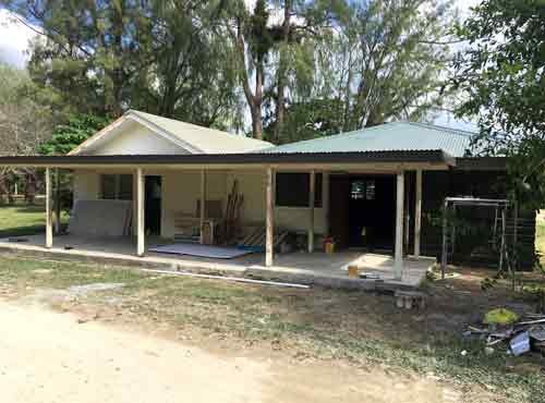 Exterior view of a single-story house with a covered patio and tropical trees.