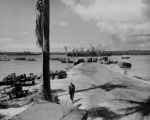 WWII wharves with ships, trucks, barrels, and a soldier on a tropical island.