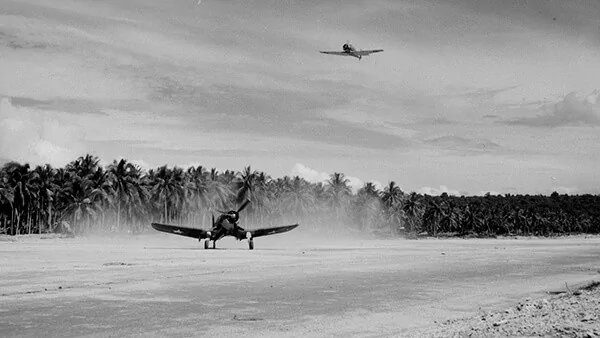 WWII fighter plane landing on a sandy airstrip with palm trees and another plane in the sky.