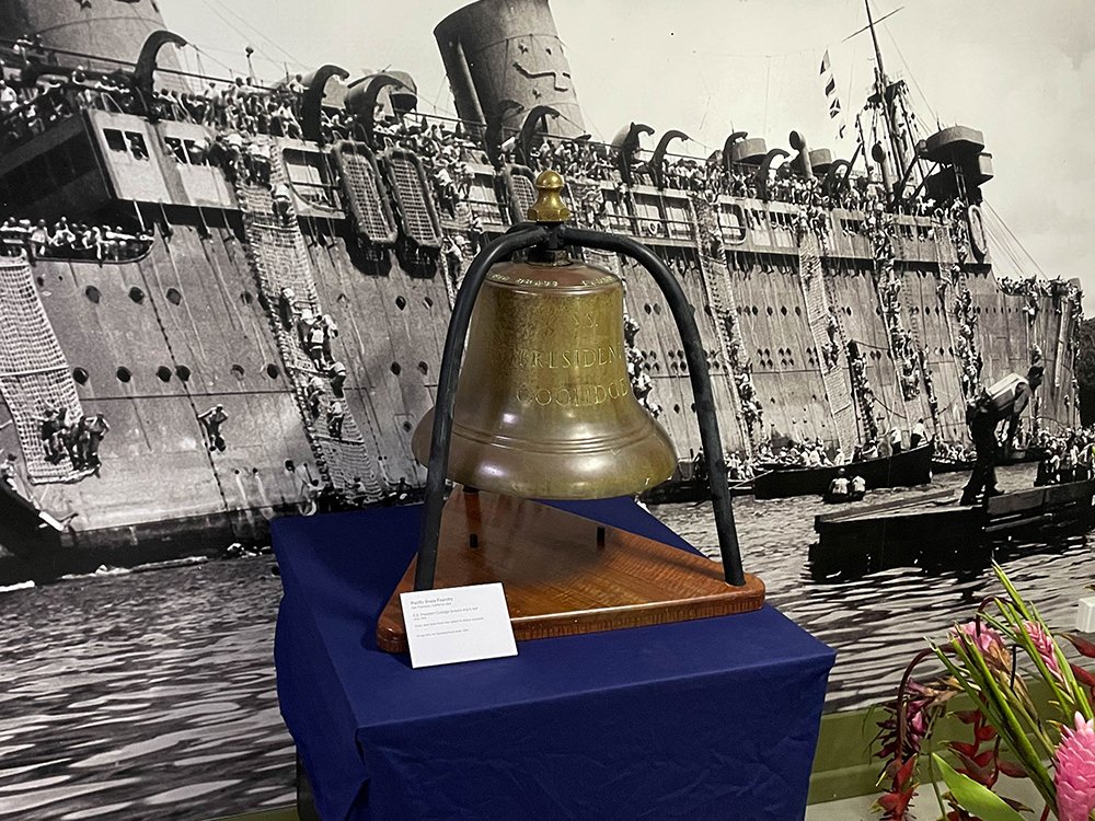 Bronze bell inscribed "President Coolidge" on display before a mural of a ship.