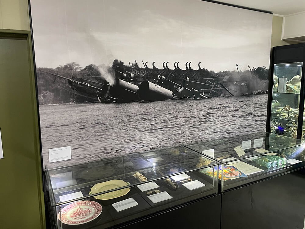Museum display with a large photo of sunken ships and artifacts in glass cases.