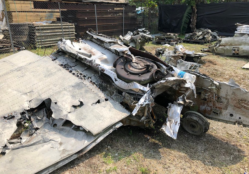 Wreckage of a damaged airplane wing and fuselage in an outdoor salvage yard.