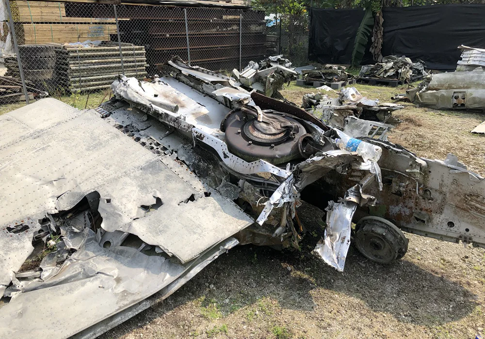 Wreckage of a damaged airplane wing and fuselage in an outdoor salvage yard.