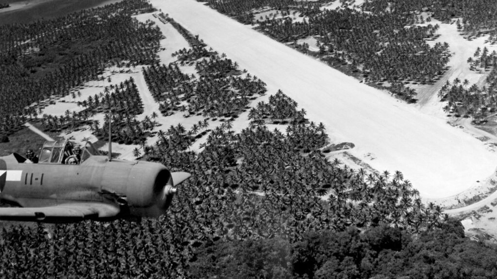 WWII aircraft flies over a tropical airfield with palm trees