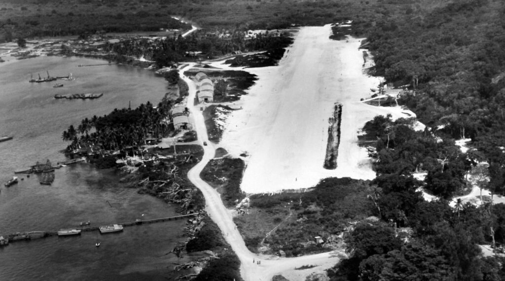 Aerial view of a wartime airfield with hangars and a runway on a tropical island.
