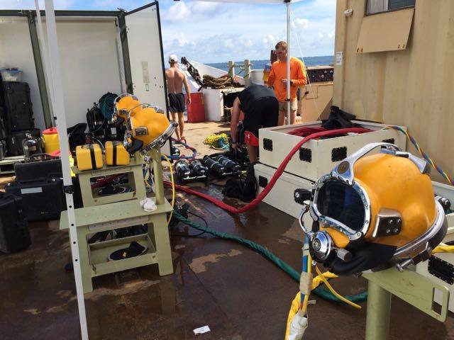 Divers prepare diving equipment and helmets on a boat deck.