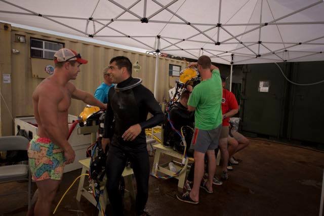 Divers prepare for a dive, with one in a full diving suit and helmet.
