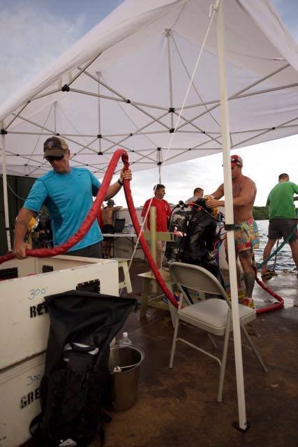 Divers prepare gear under a tent, including a scuba tank and red hoses.