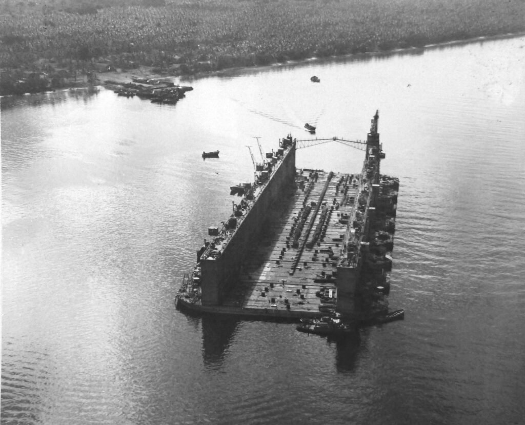 Aerial view of a large floating dry dock in a river, with ships and a lush green shoreline.