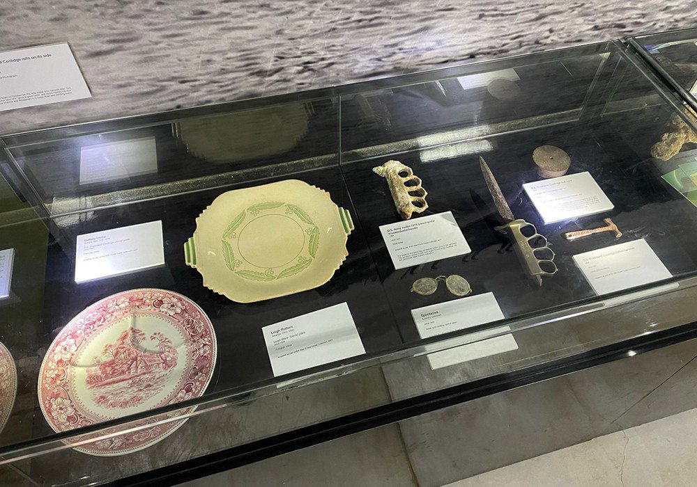Museum display case with historical artifacts: plates, brass knuckles, knife, and coins.