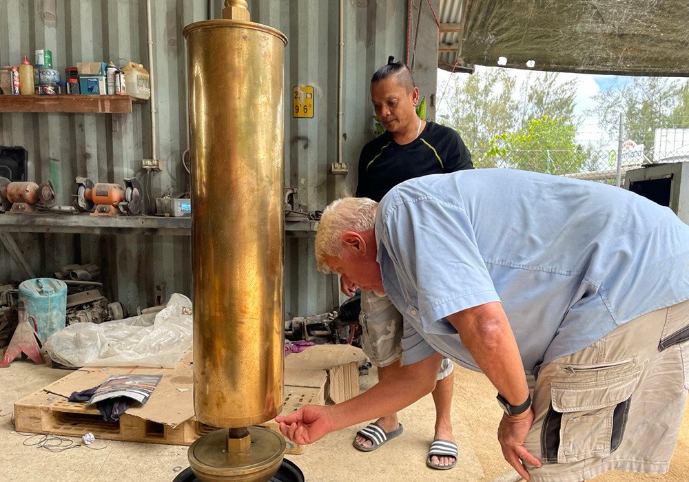 Two men examine a large brass cylinder in a workshop setting.
