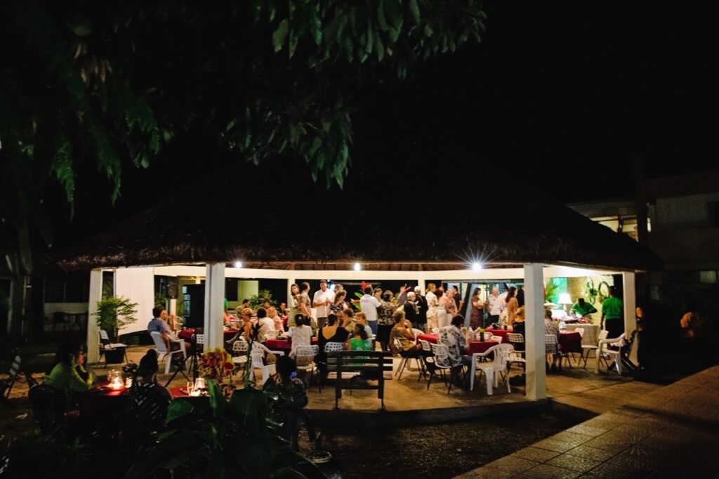 People dining and socializing at an outdoor evening event under a thatched roof.
