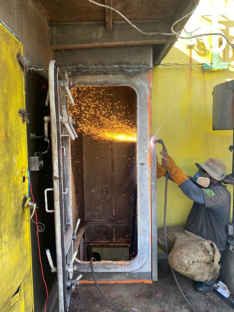 Welder in protective gear cutting metal with a plasma torch near a large industrial door.