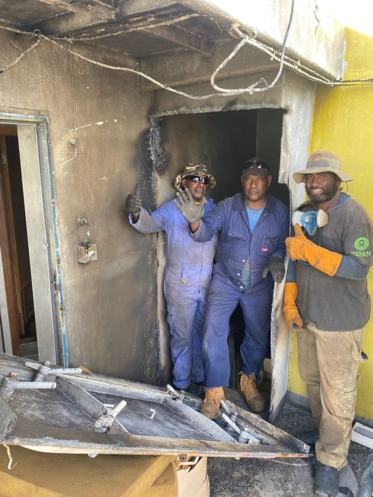 Three men in work clothes stand in a damaged doorway, one giving a thumbs up.