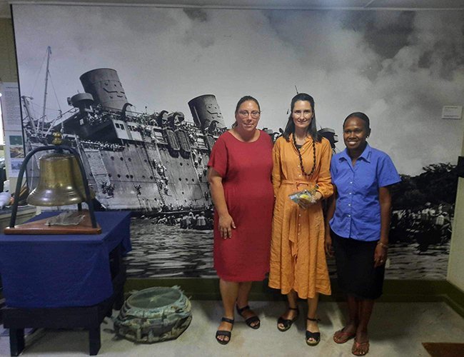 Three women stand before a large black and white mural of a shipwreck.