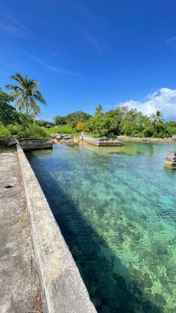 Crystal clear turquoise water in a shallow inlet with concrete walls and lush green vegetation under a blue sky.