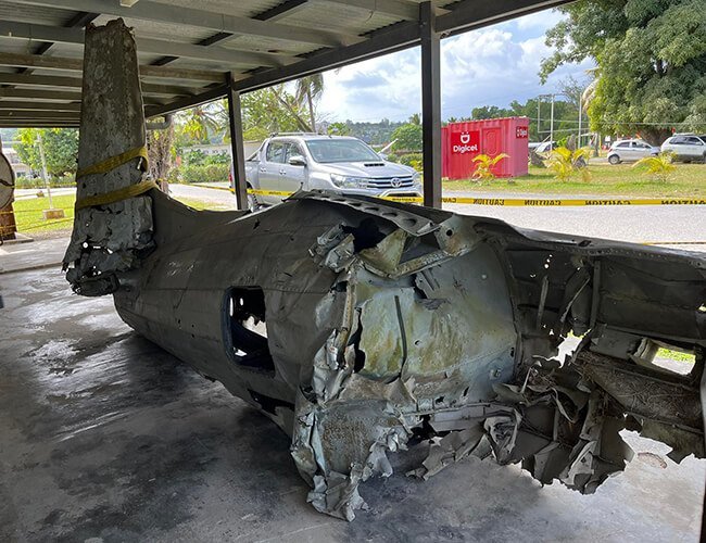 Damaged airplane wreckage under a shelter, Digicel container in background
