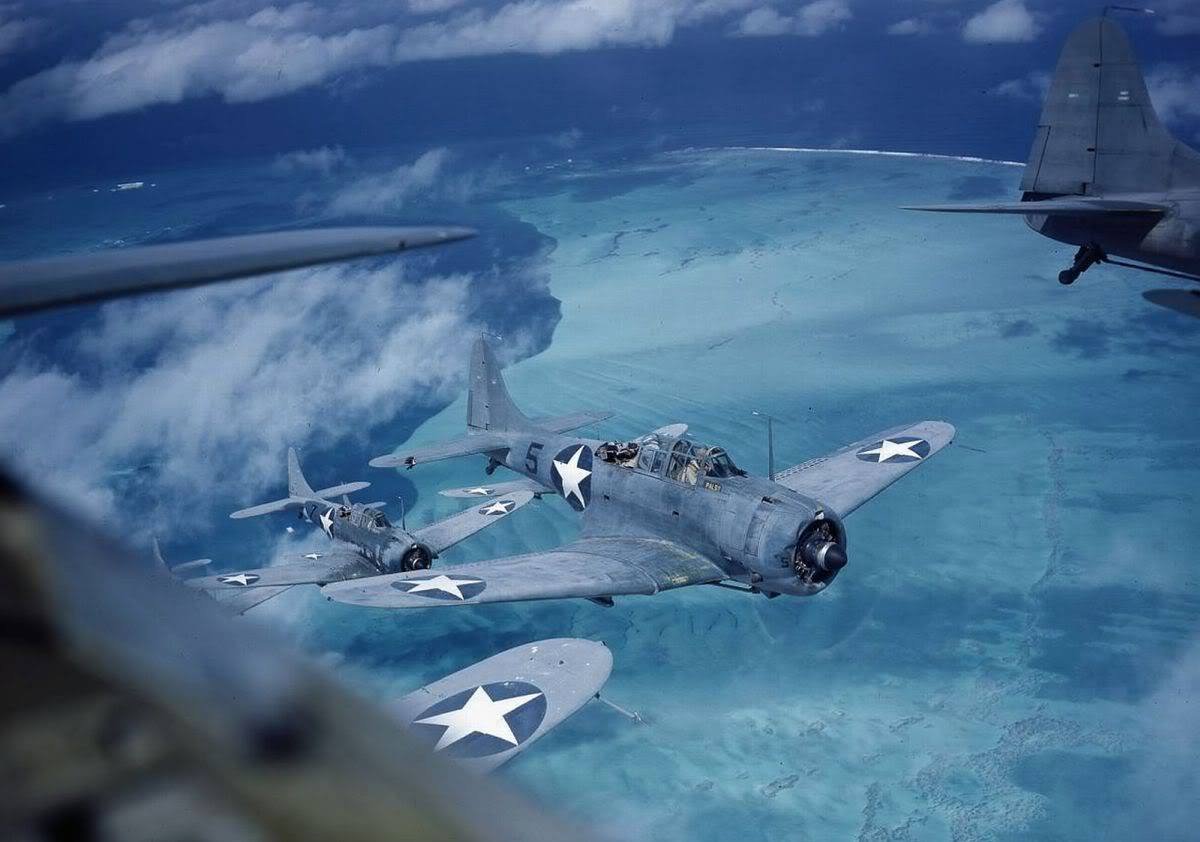 US Navy dive bombers flying over a tropical ocean, part of an airfield formation.