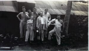 Group of six men in casual attire posing outdoors in front of a corrugated metal building.