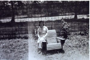 Woman in 1940s attire sits on a wooden bench in a garden.