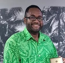 Lengkon Tokon smiles in a green patterned shirt, holding a plaque.
