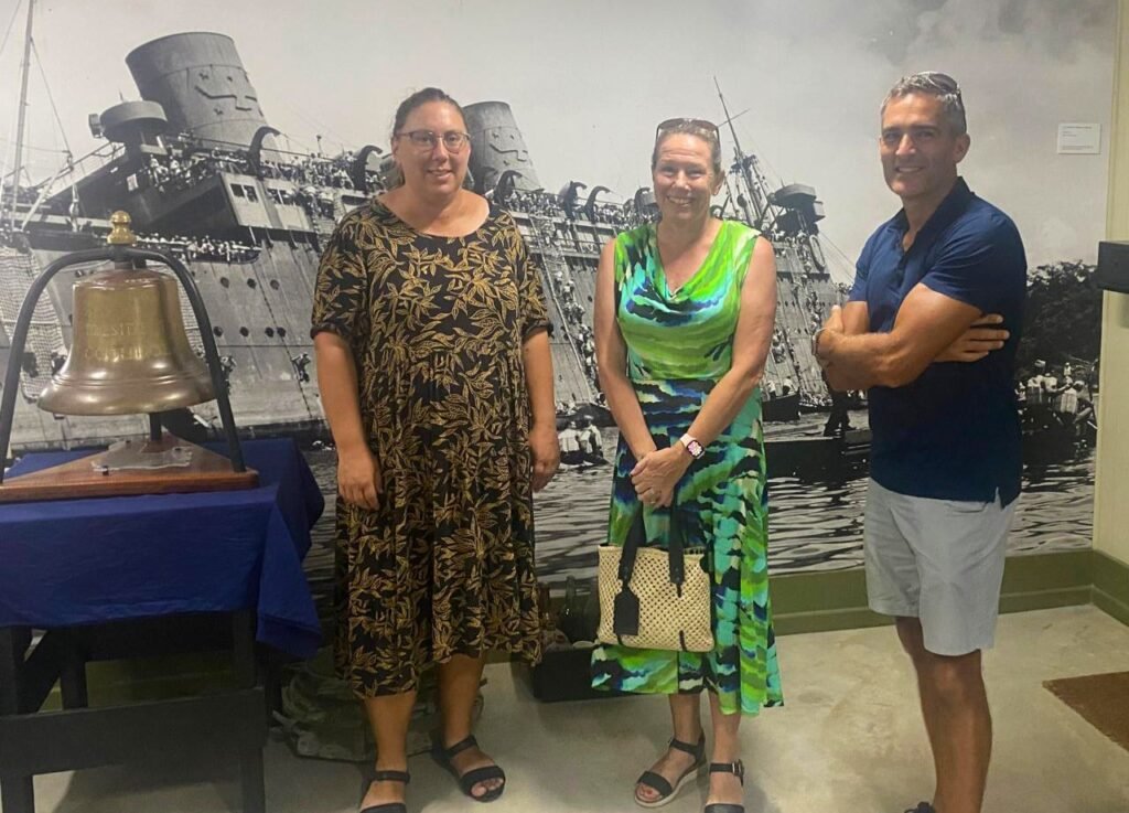 Three people stand before a historical ship mural and a ship's bell.