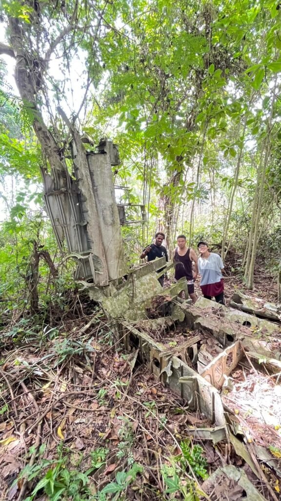 Wreckage of an aircraft in a dense jungle with three people standing nearby.
