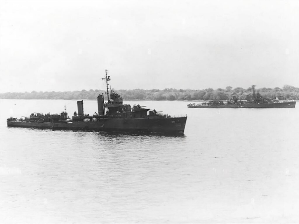 Two naval destroyers sail on calm water with a tree-lined shore in the background.